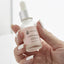 Tiny clear bottle with pink label and medicine dropper cap on white background. Pink label with black lettering reads " Yegi Beauty Super Bonder Accelerate Curing Maximize Retention" held in hand by woman with white finger nails.